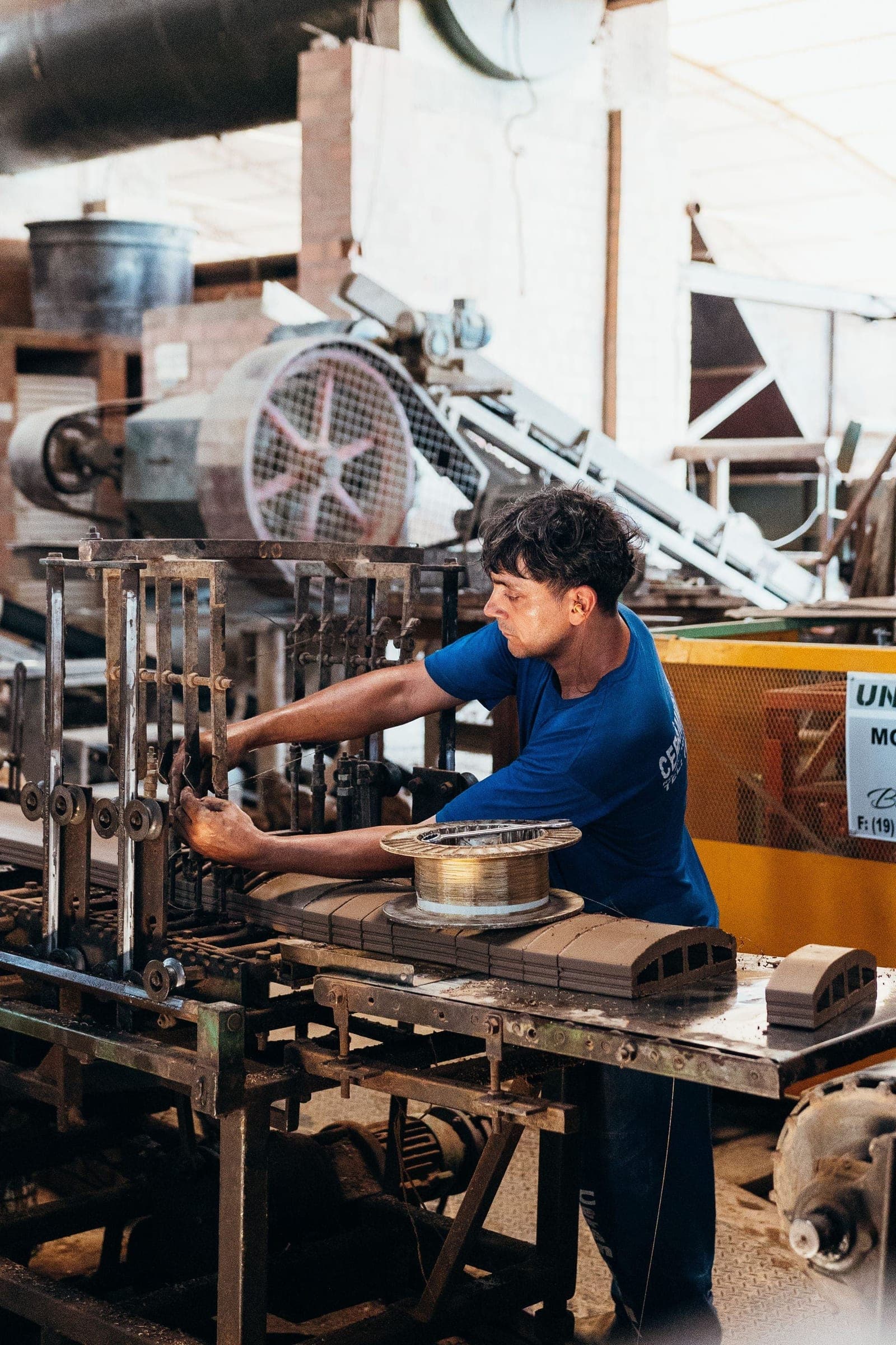 Worker in a blue shirt adjusting wire-forming machinery beside a spool of copper wire